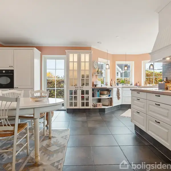 Et lyst, funktionelt køkken med hvide skabe og en central spiseplads. Til højre ses en ovn indbygget i et skab. Store vinduer giver naturligt lys og udsigt til en have.