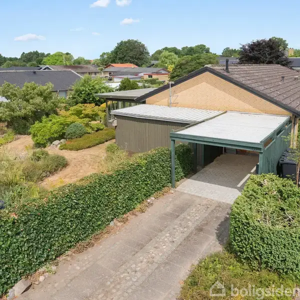 Et én-etagers hus med garage og tilhørende have. Huset er omgivet af grøn vegetation i et beboelsesområde med flere lignende huse. Vejret er klart og solrigt.