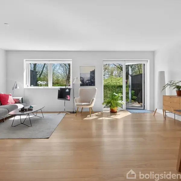 Stue med sofa og sofabord på tæppe, belyst af dagslys fra vinduer. En lænestol står nær en plante, og et maleri hænger på væggen. Trægulv og minimalistisk indretning.