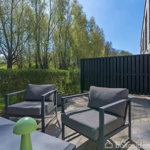 Terrassemøbler står på en belagt terrasse, omgivet af en hækbeskyttet have. I baggrunden ses høje træer under en klar blå himmel. En åben glasdør forbinder terrassen med bygningen.