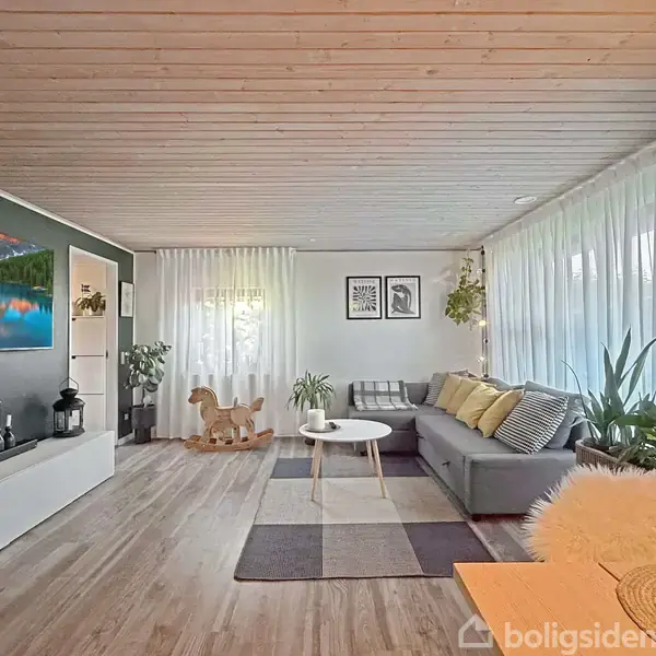 En stue med en grå sofa, dekoreret med puder, står ved et vindue med gardiner. Et rundt sofabord foran sofaen. Rummet har træloft, planter og vægkunst.