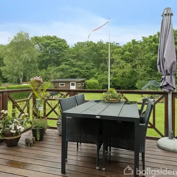 Havebord med stole står på en overdækket terrasse; omgivet af planter, parasol og udsigt til frodig have og skov. I baggrunden ses en flagstang og lille træbygning.