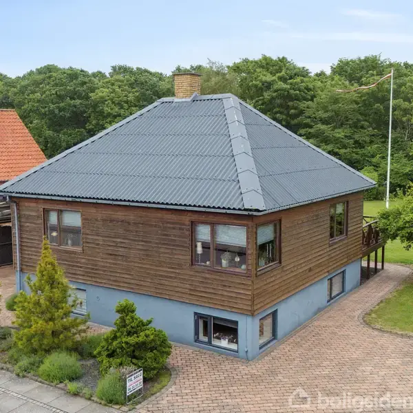 Et hus med brun træfacade står på en brostensbelagt grund med have. Omkring huset er der træer og buske. En flagstang står til højre, og en anden bygning anes i baggrunden.