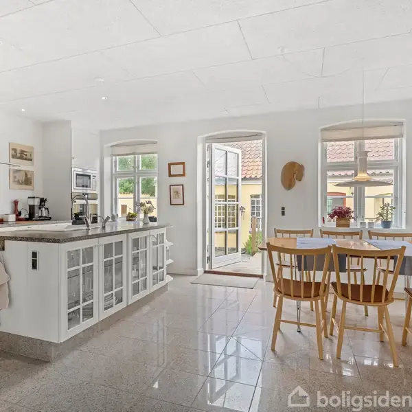 Køkkenet har et stort køkkenbord med hvide skabe, kaffemaskine og dekorationer. Der er tre træstole ved et bord. Sollys strømmer ind gennem store vinduer og en åben dør.