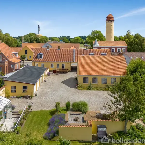 Gårdhus med gule mure og rødt tegltag står omkring en grusplads. En fransk have med blomsterbede og et drivhus ligger foran, mens et rundt vandtårn ses i baggrunden.