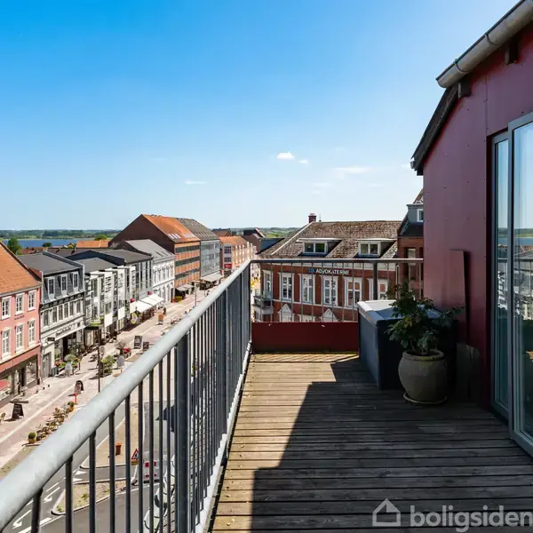 Balkonen strækker sig ud fra en bygning med glasdør, omgivet af en by med farverige bygninger og røde tage under en klar blå himmel.