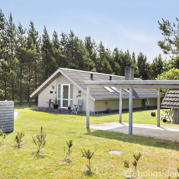 Sommerhus med gråt tag ligger fredeligt på en græsplæne omgivet af skov. Udendørs terrasse med stole og træskur i baggrunden. Ingen synlig tekst.