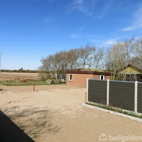 En have med sandet grund domineret af et lavt hegn og omgivende træer, med en murstensbygning i baggrunden under en klar blå himmel.