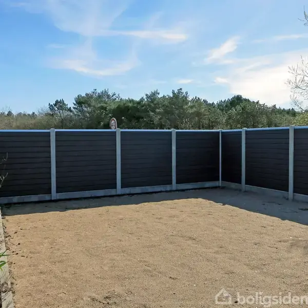 En sandet gårdhave omkranset af grå brædder og høje hegn; omgivet af høje trækroner og blå himmel.
