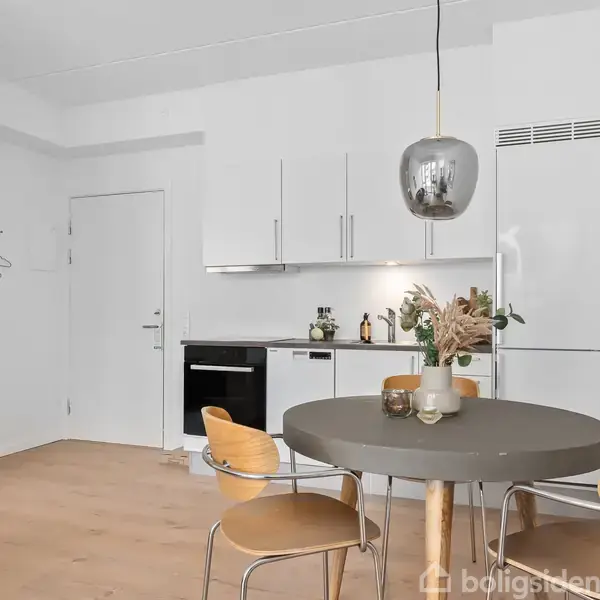 Et rundt spisebord med fire træstole står i et lyst køkken. På bordet er en vase med tørrede blomster. Baggrunden har hvide køkkenskabe, køleskab og en ovn.