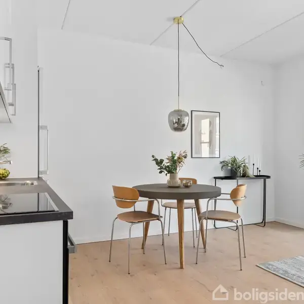 Et moderne køkken-alrum med minimalistisk design. Runde træborde og stole står under en pendellampe. Køkkenbordet har sorte overflader. Hvide vægge og store vinduer skaber et lyst rum. Ingen tekst findes.