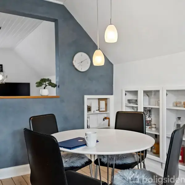 Rundt bord med sorte stole står i en spisestue med blå væg. En åben hylde viser spiritusflasker. Hængelamper tilfører lys, mens et ur hænger over en vinduesåbning mod en anden stue.