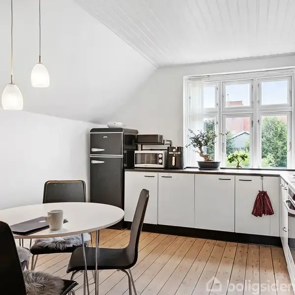 Et lyst køkken med hvide skabe og sorte håndtag. Et rundt bord med sorte stole står midt i rummet. Kælkelys hænger fra loftet, og et vindue viser en have udenfor.