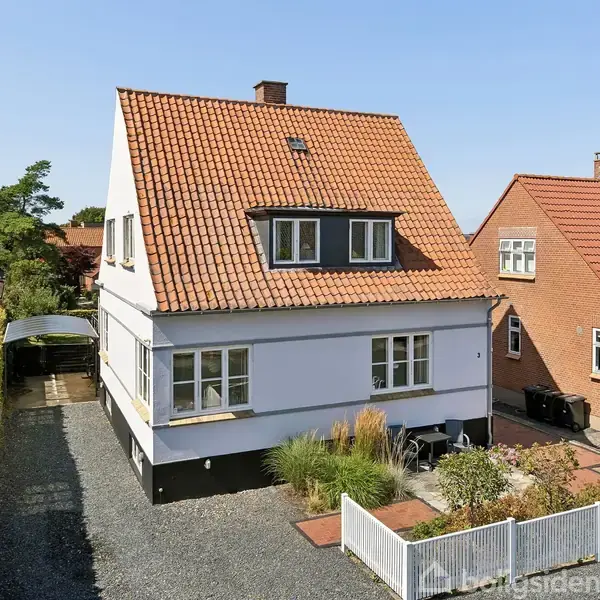 Hvidt, to-etagers hus med rødt tegltag og kvistvinduer, omgivet af grusbelagt indkørsel og have med grønne planter. Nabohus til højre er rødt med et lignende tag.