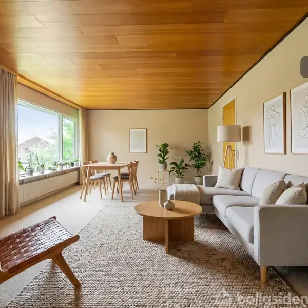Stue med moderne indretning; sofa og loungestol står på en tæppebelagt gulv, med et rundt træbord. Store vinduer og spisebord i baggrunden. Tekst: "Visualisering".