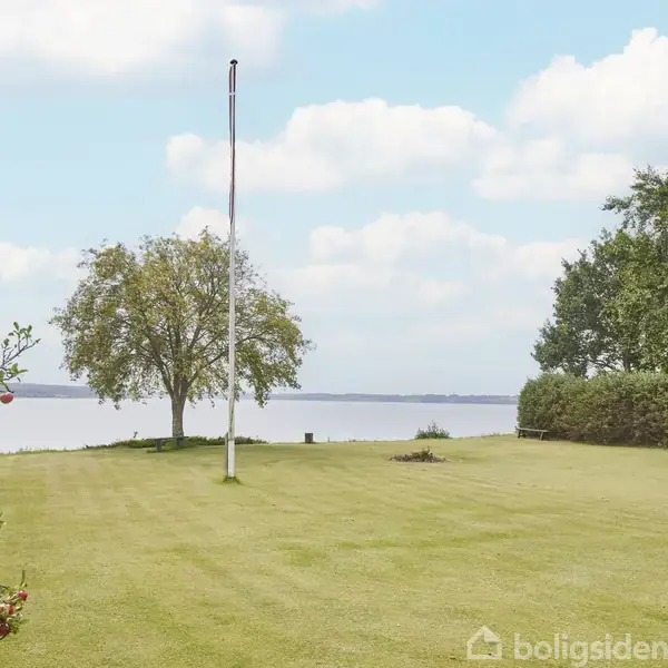 Flagstang står ubenyttet på velholdt græsplæne ved æbletræ, med udsigt over stor sø. Omgivet af buske og træer under blå himmel med spredte skyer. Udsigt fremhæver naturlig skønhed.