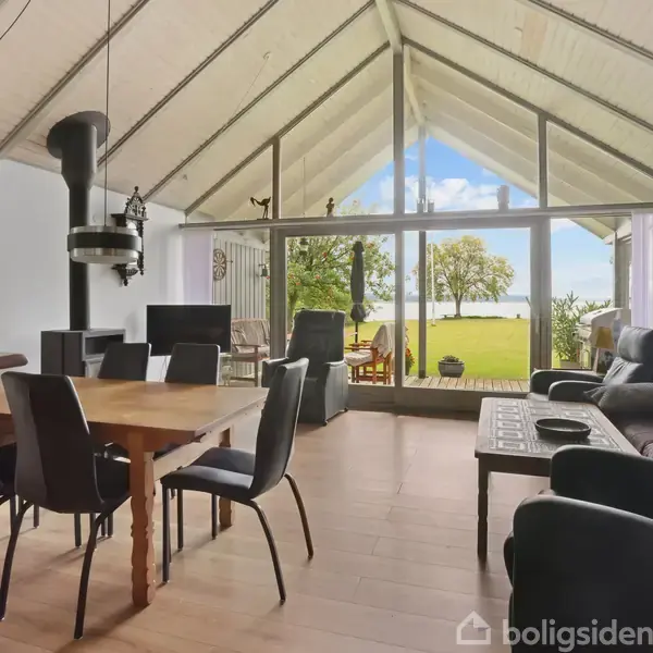 Stue med spisebord og stole står ved siden af en sofa og sofabord. Store glasvinduer åbner ud mod en have med udsigt til græsplæne og træer i baggrunden.