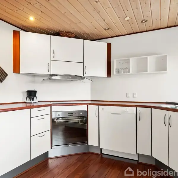 Et hvidt, moderne køkken med træbordplader og loft, opvaskemaskine og ovn indbygget. En kaffemaskine står på bordet, og grydelapper hænger på væggen. Rummet har trægulv og loftspotlys.