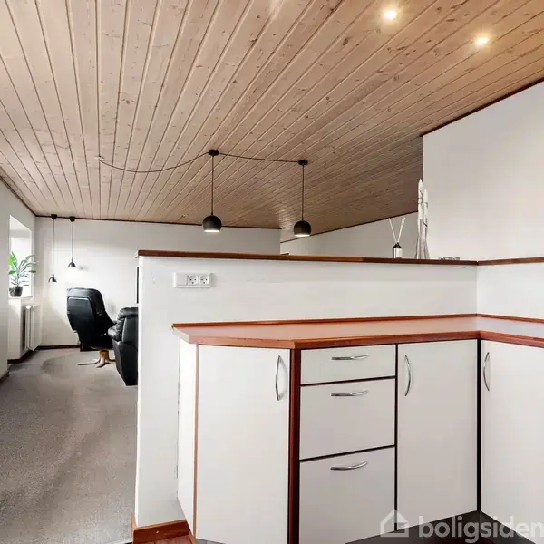 Køkken med hvidt skab og træbordplade fører til en stue med træloft og sort lænestol. Indirekte lys og minimalistisk pynt skaber en lys og stilfuld atmosfære.