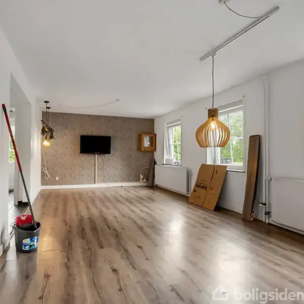 Rummeligt lokale med trægulv og hvide vægge, fladskærms-tv på væggen, pendellampe i træ, malingforsyninger og værktøj i hjørne, to vinduer giver udsigt til en grøn baggrund.