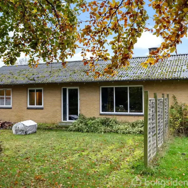 Et gult murstenshus med store vinduer og sort tag står i en have med græsplæne. Høstfarvede træer omkranser huset, og en stak brænde ligger ved siden af.