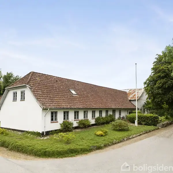 Et hvidt, stråtækt hus står ved en vej og er omgivet af grønne buske og træer. Huset har røde tegl på taget, og himlen er klar.