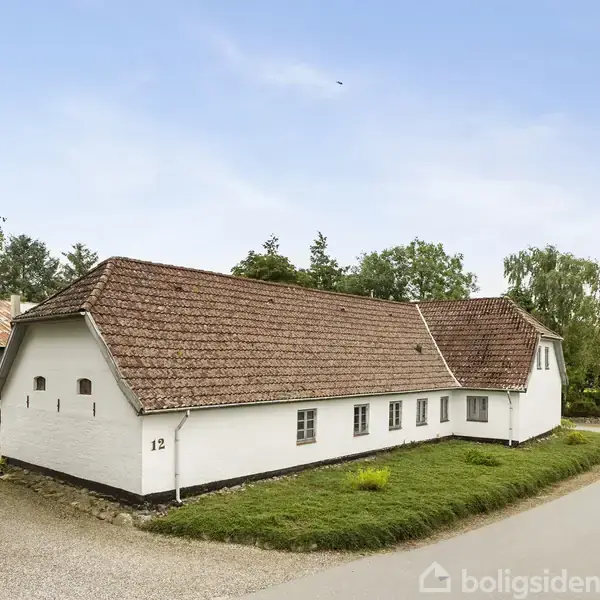 En lang, hvid murstensbygning med rødt tegltag står ved en grusvej. Træer omgiver huset, der har nummeret 12 på siden og ligger tæt på en asfalteret vej.