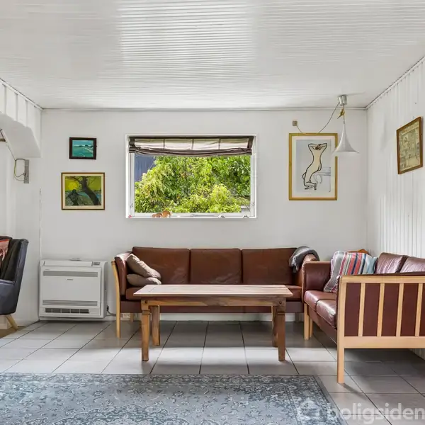 Stue med en brun lædersofa og sofabord står på et tæppe. Vinduet viser grønne omgivelser udenfor. Væggene er dekoreret med kunst, og et par stole står ved siden af.
