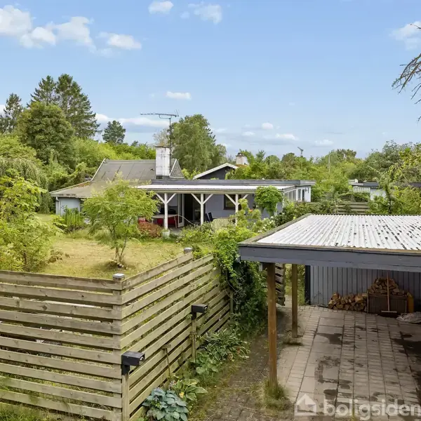 Et hus står stille i en frodig have, omgivet af træer. En carport ses til højre, delvist dækket af planter, med brændestakke i baggrunden under halvtaget.