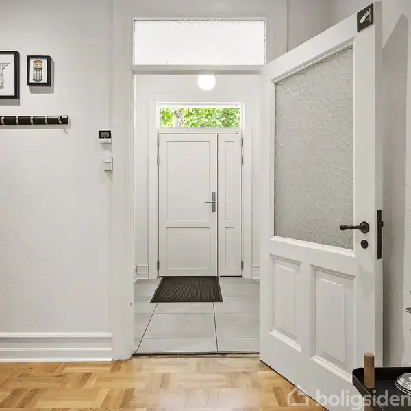 Dør står åben i en hvidmalet entré med parketgulv. Væggene er dekoreret med billeder, og to grønne stearinlys står på et bord. Yderdør med vindue ses i baggrunden.