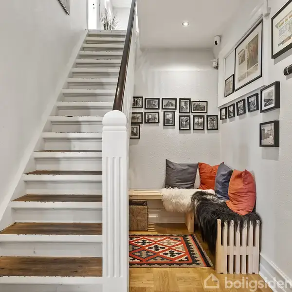 Trappe til venstre fører opad; ved siden af er en hyggekrog med farverige puder og tæppe. Væggene har mange indrammede billeder, der skaber en hjemlig atmosfære.