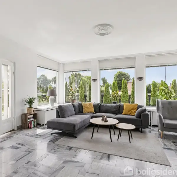 En grå sofa med gule puder står ved vinduer og omkringliggende naturelementer. Stuen har marmorgulv og åbner op til en terrasse gennem glasdøre, med moderne og minimalistisk stil.