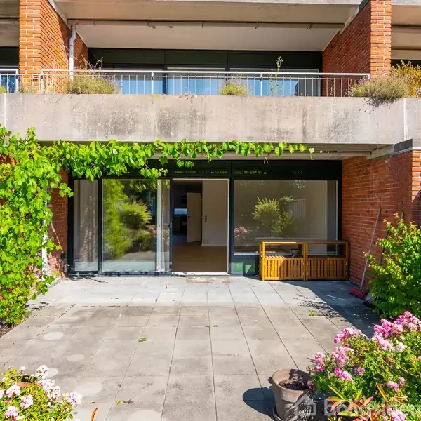 Terrassen har fliser, omgivet af blomster og planter. En stor glasdør åbner ind til huset. Røde murstensvægge og en overhængende balkon med planter udgør bygningens struktur.