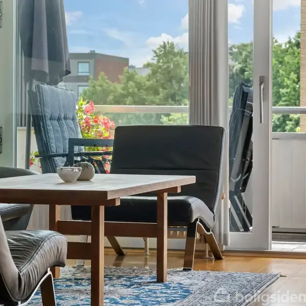 Et bord med stole står i en stue med åben altandør. Udenfor ses en balkon med blomster og grønt. Sollys skinner ind gennem døren.