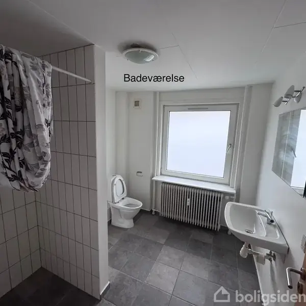 Badeværelse med hvide fliser viser en brusekabine til venstre, toilet midt i rummet, og en håndvask med spejl til højre. Et stort vindue skaber naturligt lys. Tekst: "Badeværelse".