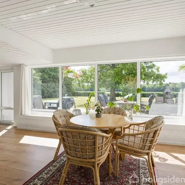 Rundt spisebord med flettede stole står på tæppe i lys stue. Store vinduer viser terrasse og grøn have. Koselig og indbydende atmosfære med naturligt lys.