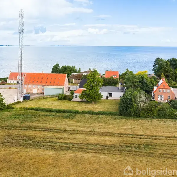 Markerede bygninger med røde tage står stille omgivet af frodig vegetation ved kysten. I baggrunden strækker havet sig til horisonten, under en blå og skyet himmel.