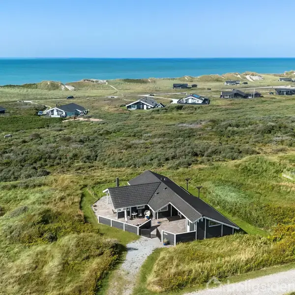 Et hus med mørkt tag ligger fredeligt blandt sandklitter og græs. Flere lignende huse ses spredt over landskabet. I baggrunden skimtes den klare, blå himmel og havet.