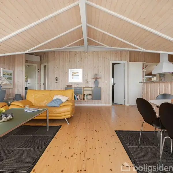 Gule sofaer står i en stue med trævægge og tagbjælker; et bord og stole ses ved siden af, med en åben køkkenafdeling i baggrunden.