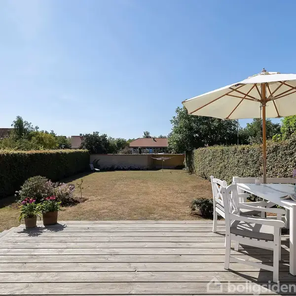 Terrasse med hvidt møbelsæt og stor parasol. Omgivet af grøn hæk, fører ud til tør græsplæne med blomsterkrukker. Klar, blå himmel i baggrunden.