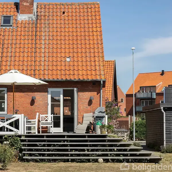 Et rødt murstenshus med rødt tegltag har en terrasse med møbler og en parasol. Det er omgivet af en have, og der er nabobygninger i baggrunden.