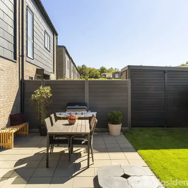 Terrassebord med stole står ude på en flisebelagt terrasse. En grill står ved et hegn, omgivet af planter. Græsplæne og buske ses i baggrunden under en klar himmel.