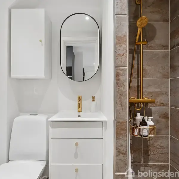 Toilet, håndvask med ovalt spejl og gyldne armaturer står i et moderne badeværelse med brune fliser i bruseområdet. Vægmonteret skab over toilettet.