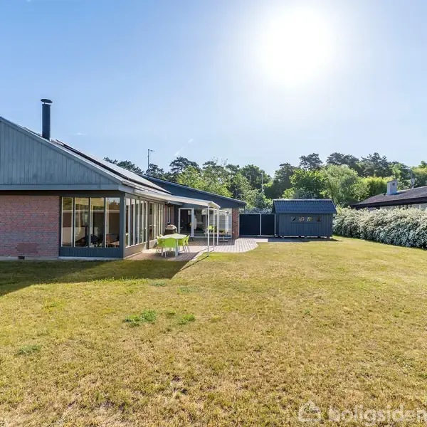 To liggestole står på en flisebelagt terrasse ved siden af et hus med en overdækket del, omkranset af en grønt klippet græsplæne og en høj hæk under en klar blå himmel.