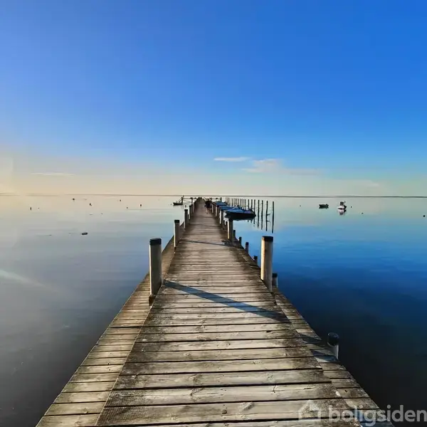 En lang, træmole strækker sig roligt ud over en spejlblank fjord under en klar blå himmel, omgivet af spredte både og bøjer i vandet.
