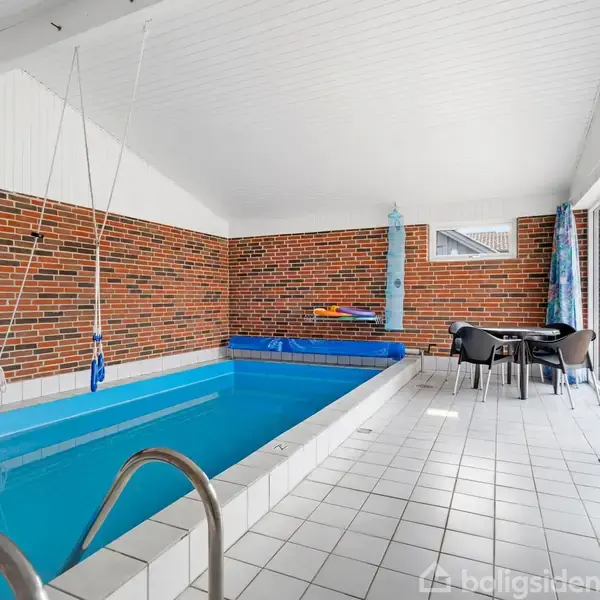 Et moderne hus med store vinduer står på en velplejet græsplæne. Huset har en terrasse med borde og stole og omgives af træer og beskærmende hegn.