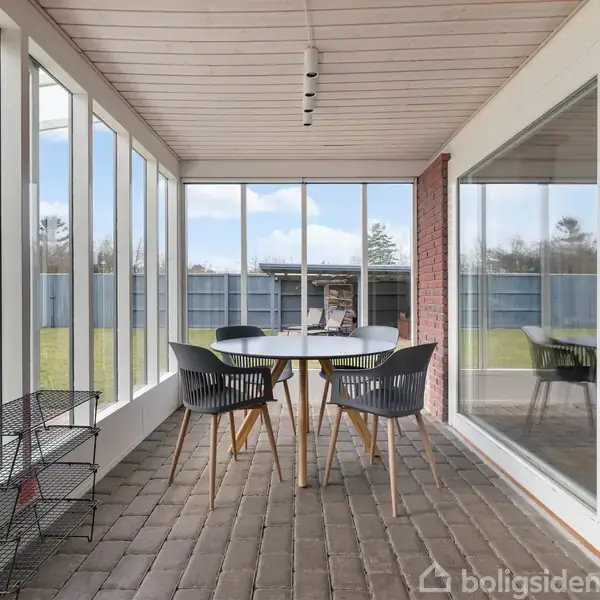 Rundt bord omgivet af fire stole står i en lys havestue med store vinduer. Udenfor ses en græsplæne og et hegn. En reol står ved væggen.