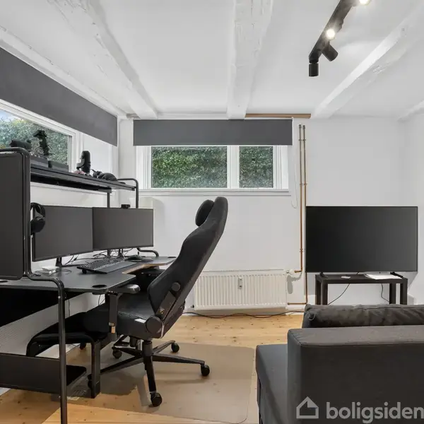 Kontorstol foran et skrivebord med to computerskærme; ved siden af en grå sofa og en stor fladskærm. Rummet har hvide vægge og et trægulv, med store vinduer til venstre.