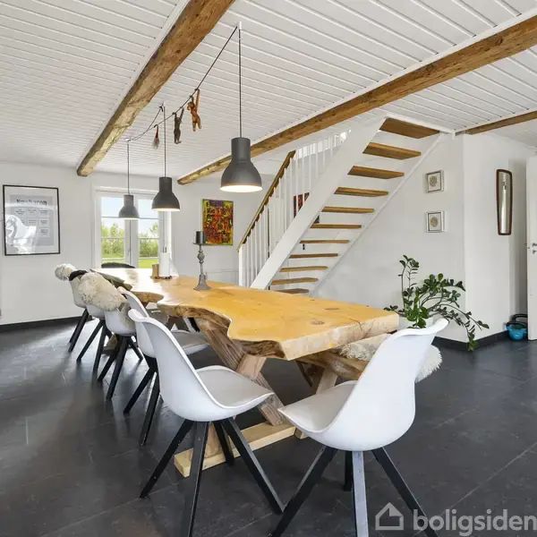 Et stort træbord omgivet af hvide stole står centralt i et moderne rum med loftsbjælker. I baggrunden ses en trappe, planter og en tv-skærm. Vinduerne lukker lys ind.