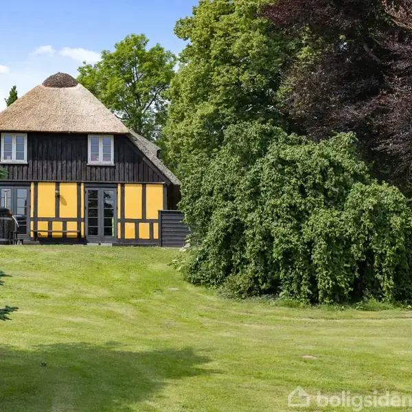 En charmerende bindingsværkshus med stråtag står på en velplejet græsplæne omgivet af tætte træer. Sollys oplyser den gule facade, og huset er omkranset af frodig vegetation.